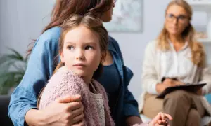 Dziewczynka w ramionach matki, zaniepokojona, szuka wsparcia. W tle terapeuta. Obrazuje skutki niepraktykowania wiary przez rodziców dla duchowości dziecka.