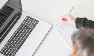 Osoba pisze w notatniku obok laptopa. Jak praktykować wiarę w miejscu pracy zachowując profesjonalizm, to temat refleksji.