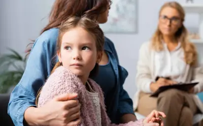Dziewczynka w ramionach matki, zaniepokojona, szuka wsparcia. W tle terapeuta. Obrazuje skutki niepraktykowania wiary przez rodziców dla duchowości dziecka.