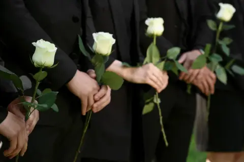 Ludzie w czerni trzymają białe róże. Mogą symbolizować żałobę, ale też nadzieję, która bywa trudna dla osób wierzących, gdy pojawiają się przyczyny lęku przed śmiercią.
