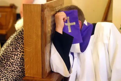 Spowiedź w konfesjonale. Kapłan w białej albie i fioletowej stule z krzyżem, zasłaniający twarz. Rozważania o skutkach nieobecności na mszy niedzielnej w świetle prawa kanonicznego.