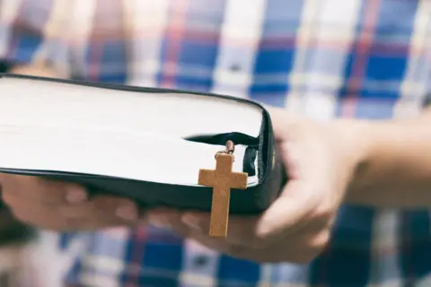 Osoba w kratę trzyma Biblię z drewnianym krzyżykiem, symbolizującym dwa przykazania miłości.