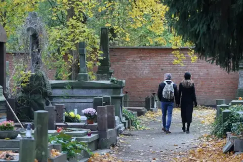 Jesienne zaduszki na cmentarzu. Para spaceruje alejką wśród nagrobków. Jak zabezpieczyć wiązanki i znicze przed kradzieżą?