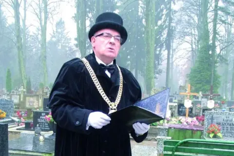 Mistrz ceremonii w czarnym stroju i z łańcuchem na szyi prowadzi pogrzeb świecki, podkreślając różnicę między nim a wyznaniowym.