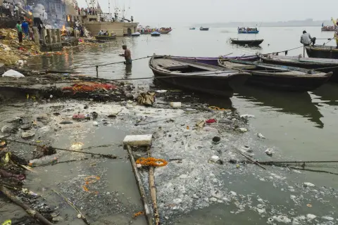 Rzeka Ganges, święta w hinduizmie, jest miejscem rytuałów i kremacji. Widać łodzie i ludzi na brzegu, ale też śmieci unoszące się na wodzie.