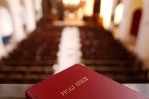 Czerwona Biblia leży na pierwszym planie, w tle widać rzędy pustych ławek w kościele. Szukasz odpowiedzi, jak przejść na protestantyzm?