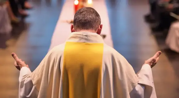Kapłan w białej szacie z żółtym pasem, z rękami uniesionymi w geście modlitwy, zastanawia się, czy praca w niedziele i święta jest zawsze grzechem. Dylemat zawodowy.