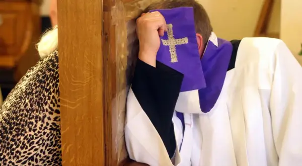 Spowiedź w konfesjonale. Kapłan w białej albie i fioletowej stule z krzyżem, zasłaniający twarz. Rozważania o skutkach nieobecności na mszy niedzielnej w świetle prawa kanonicznego.