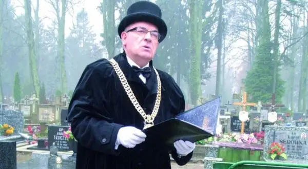Mistrz ceremonii w czarnym stroju i z łańcuchem na szyi prowadzi pogrzeb świecki, podkreślając różnicę między nim a wyznaniowym.