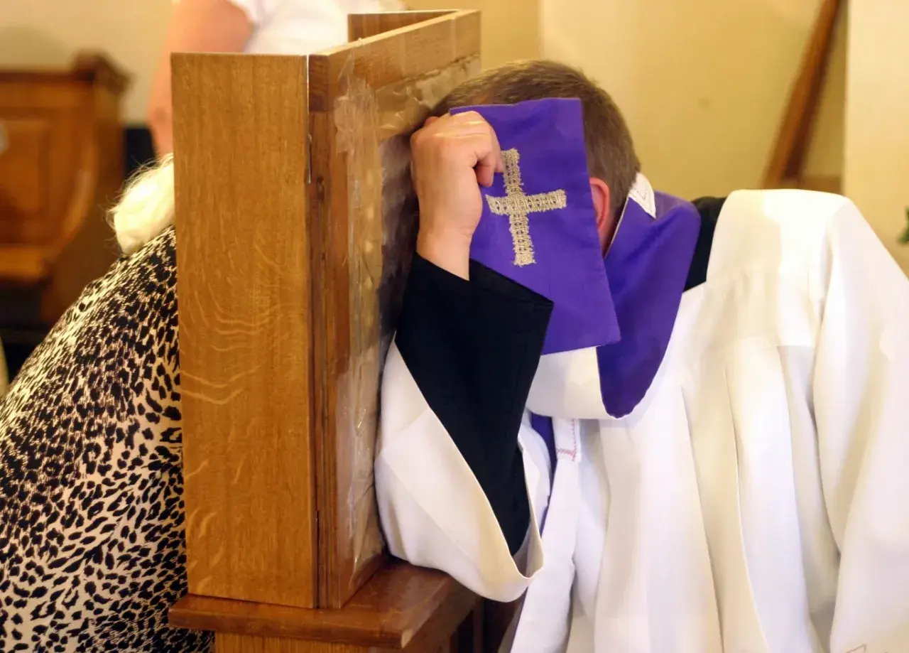 Spowiedź w konfesjonale. Kapłan w białej albie i fioletowej stule z krzyżem, zasłaniający twarz. Rozważania o skutkach nieobecności na mszy niedzielnej w świetle prawa kanonicznego.
