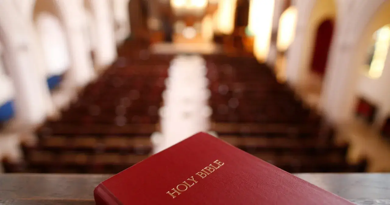 Czerwona Biblia leży na pierwszym planie, w tle widać rzędy pustych ławek w kościele. Szukasz odpowiedzi, jak przejść na protestantyzm?