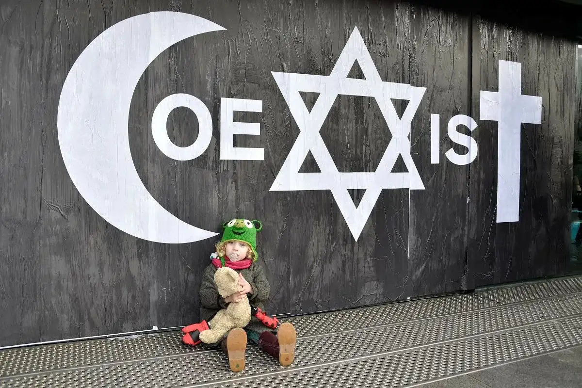 Dziecko z pluszakiem pod symbolem "COEXIST" na tle ścian, ilustrujące skutki nadmiernego intelektualizowania wiary kosztem doświadczenia.