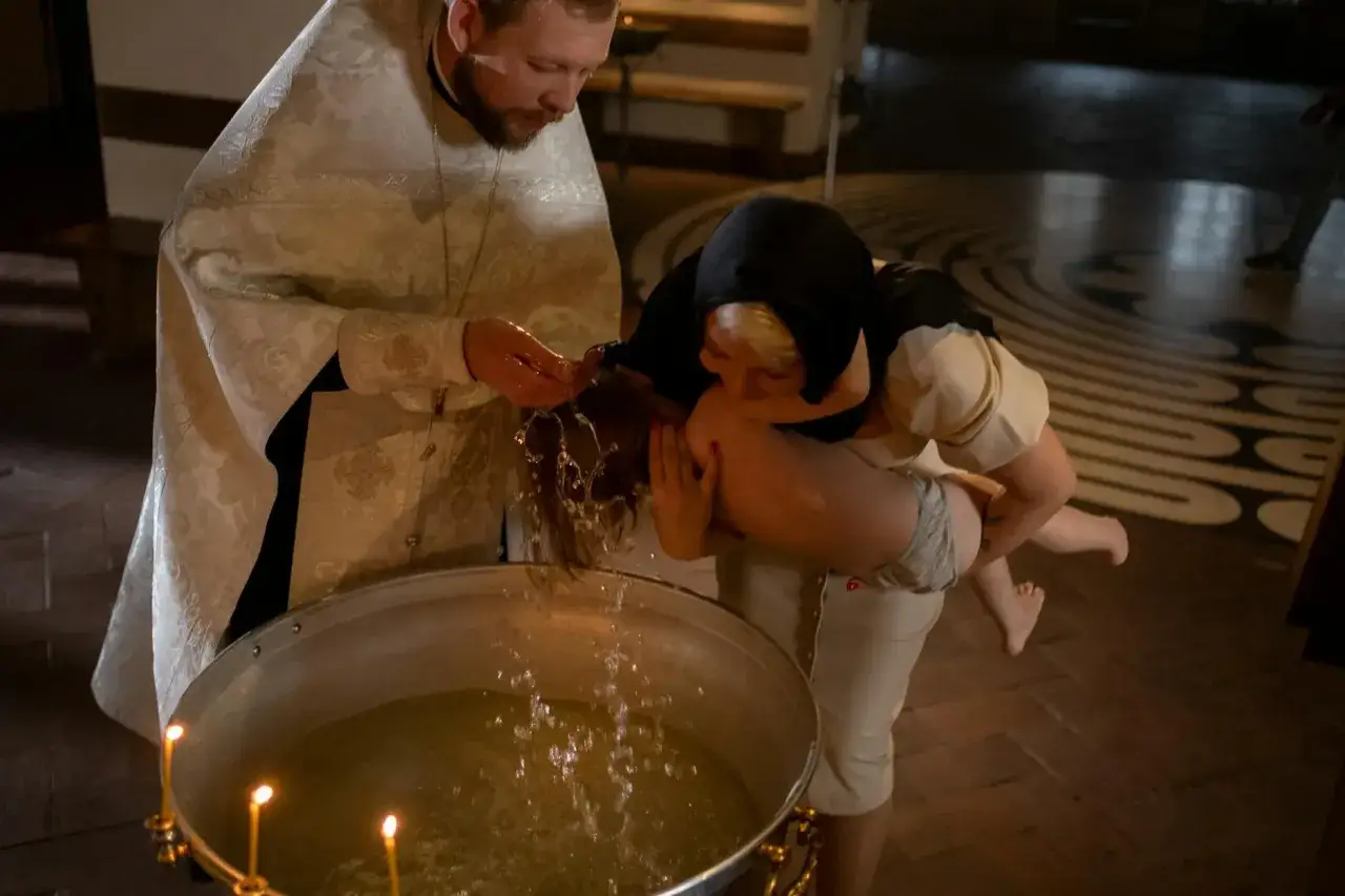Kapłan w bogatej szacie błogosławi wodę, polewając nią głowę dziecka. Matka trzyma niemowlę nad chrzcielnicą, podczas ceremonii jak poświęcić wodę.