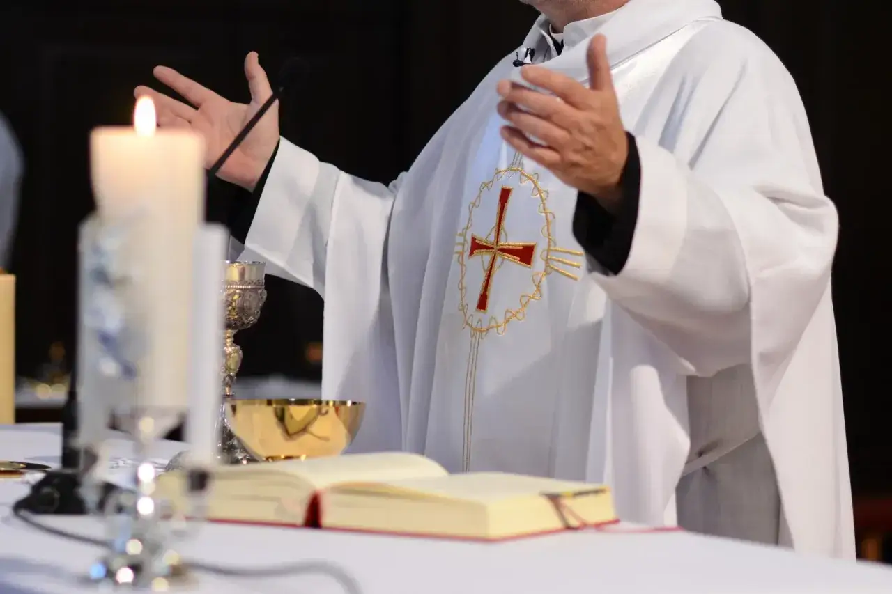 Kapłan w bieli z krzyżem na piersi, gestykuluje. Obok świeca, kielich i otwarta księga. Warto pamiętać, jakie są prawa wiernych w kontakcie z hierarchią kościelną.