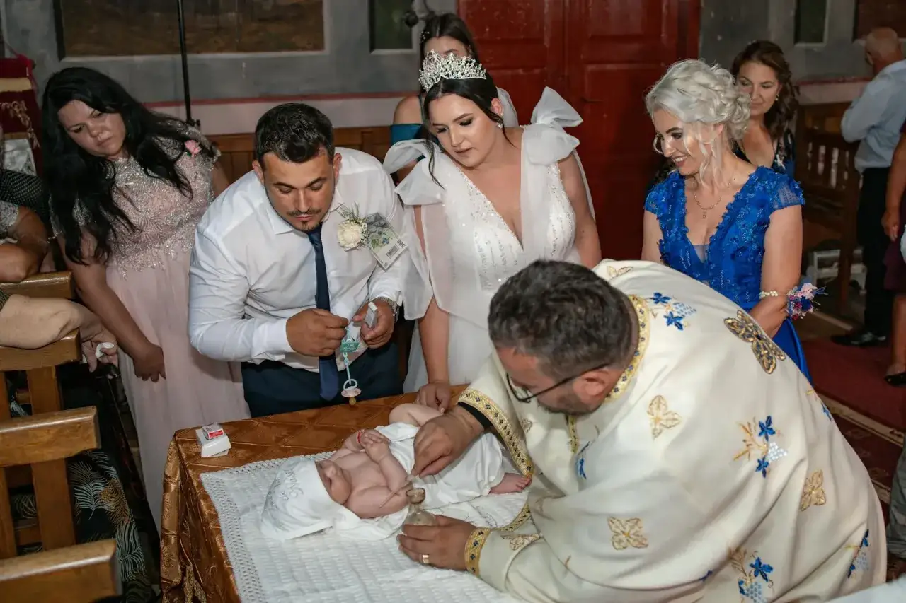 Uroczystość w kościele: kapłan w bogato zdobionej szacie jak poświęcić wodę nad niemowlęciem. Rodzice i goście patrzą z uwagą.