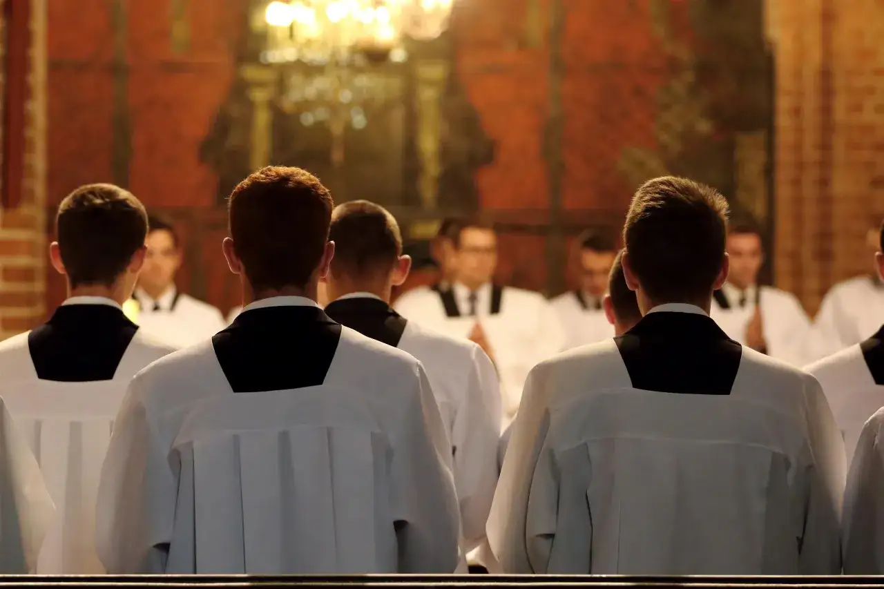 Młodzi mężczyźni w sutannach, być może klerycy, siedzą w kościele. Obraz skłania do refleksji nad przyczynami spadku powołań kapłańskich, analizy i statystyki.