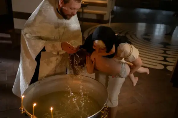 Kapłan w białej szacie błogosławi wodę, polewając nią głowę dziecka podczas chrztu.