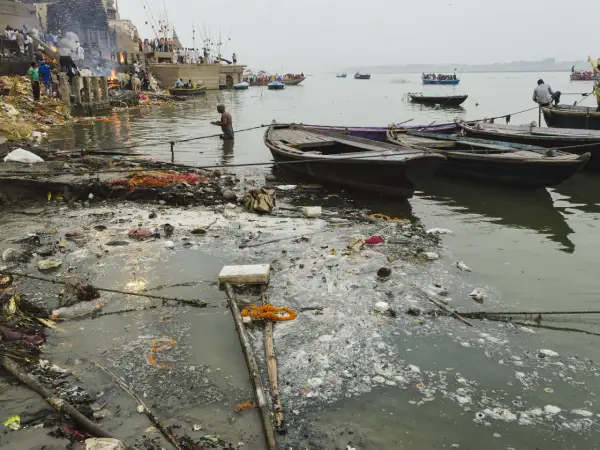 Rzeka Ganges, święta w hinduizmie, jest miejscem rytuałów i kremacji. Widać łodzie i ludzi na brzegu, ale też śmieci unoszące się na wodzie.