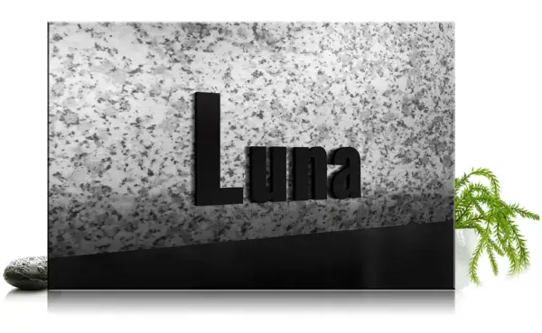 Czarna tablica z napisem "Luna" na tle granitu, z kamieniem i rośliną.