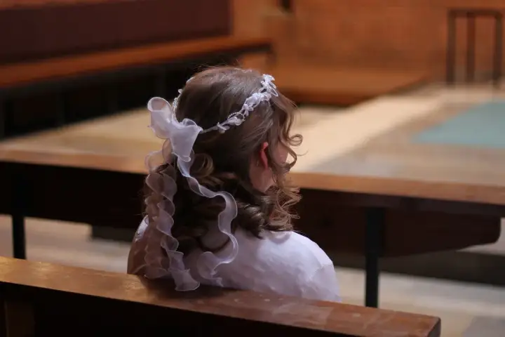 Jak rozmawiać z dziećmi o bogu i kościele porady pedagogów. Dziewczynka w białej sukience i wianku na głowie siedzi w kościele.